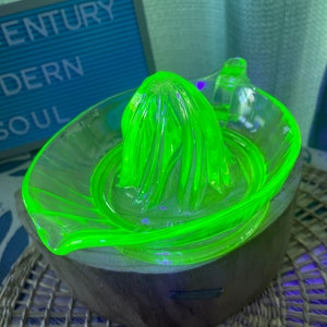 May include: A bright green glass citrus juicer with a fluted design sits on a wooden base. The juicer is made of uranium glass and glows under a black light.