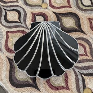 May include: A black, shell-shaped decorative object with silver accents. The shell design has radiating lines and a glossy finish. The object is placed on patterned fabric.