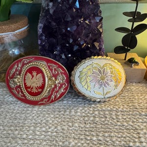May include: Two vintage belt buckles. One is red with a gold eagle and crown design. The other is silver with a gold flower design.