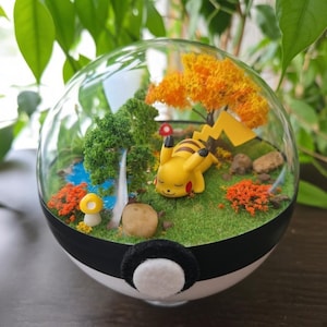 Op de afbeelding: Een bolvormig glazen terrarium in de vorm van een wit-zwarte bal met een gele Pikachu-figuur op groen mos. De scène bevat een kleine blauwe vijver, een gele paddenstoel, een groene boom, een oranje boom en kleine stenen.