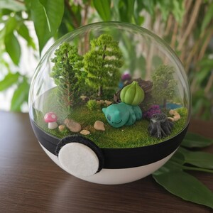 Op de afbeelding: Een bolvormig terrarium in de vorm van een wit-zwarte bal met een miniatuur boslandschap. Binnenin rust een klein groen wezen met een bolvormige plant op de rug op groen mos, samen met kleine bomen, een paddenstoel en een boomstronk.