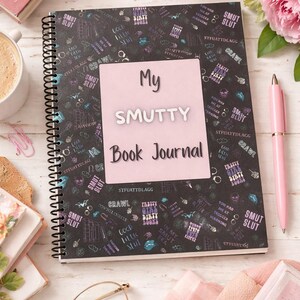 May include: A spiral-bound notebook with the text "My Smutty Book Journal" on a pink label, surrounded by a black background with various illustrations and text. A pink pen and a lit candle are also visible.