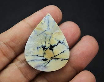 May include: A teardrop-shaped cabochon gemstone featuring a creamy off-white and pale yellow base color with distinct, dark black dendritic inclusions that resemble branching patterns or thin, jagged lines across the smooth surface.