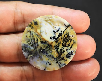 May include: A circular, flat gemstone pendant featuring a creamy white and yellow base with intricate, dark black dendritic fern-like inclusions spreading across the surface. The stone has a smooth, polished finish and a small drilled hole near the edge.