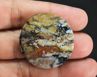 May include: A circular, polished gemstone cabochon featuring a complex, marbled pattern of earthy ochre, deep brown, creamy white, and translucent sections, accented by intricate, dark, fern-like dendritic inclusions throughout the stone surface.