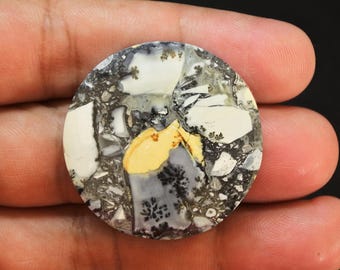 May include: A circular cabochon gemstone featuring a complex mosaic pattern of white, gray, and black fragments. The stone displays distinct dendritic fern-like inclusions in black and a prominent central patch of solid mustard yellow color.