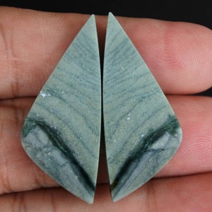 May include: Two matching, light green gemstone pieces with a triangular shape. The stones have a natural, striped pattern with darker green and white accents. The background is a dark color.