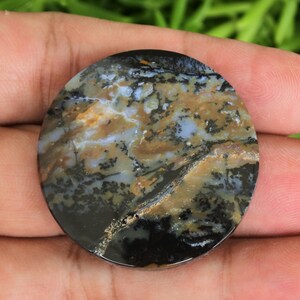 Puede incluir: Una piedra redonda y pulida con un patrón único de remolinos negros, marrones y blancos. La piedra tiene un acabado brillante y se sostiene en una mano sobre un fondo verde borroso. El diseño de la piedra se asemeja a un paisaje.