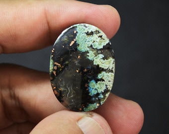May include: An oval-shaped polished gemstone featuring a dark black base with vibrant inclusions of mossy green, turquoise blue, and metallic copper-colored flecks distributed across the surface in a natural, organic pattern.
