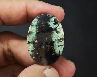 May include: An oval-shaped cabochon gemstone featuring a dark black center with mossy green and teal inclusions, accented by small flecks of metallic copper throughout the polished surface of the stone.
