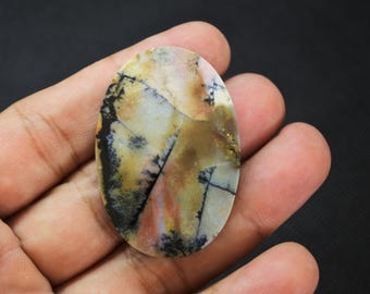 May include: An oval-shaped dendritic agate cabochon featuring a smooth, polished surface with translucent cream, tan, and brown tones, accented by intricate, dark, fern-like manganese oxide inclusions branching across the stone.