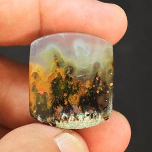 May include: A polished, shield-shaped moss agate gemstone featuring a translucent, milky white top section that transitions into a vibrant landscape of deep forest green, earthy brown, and bright orange inclusions resembling a natural forest scene.