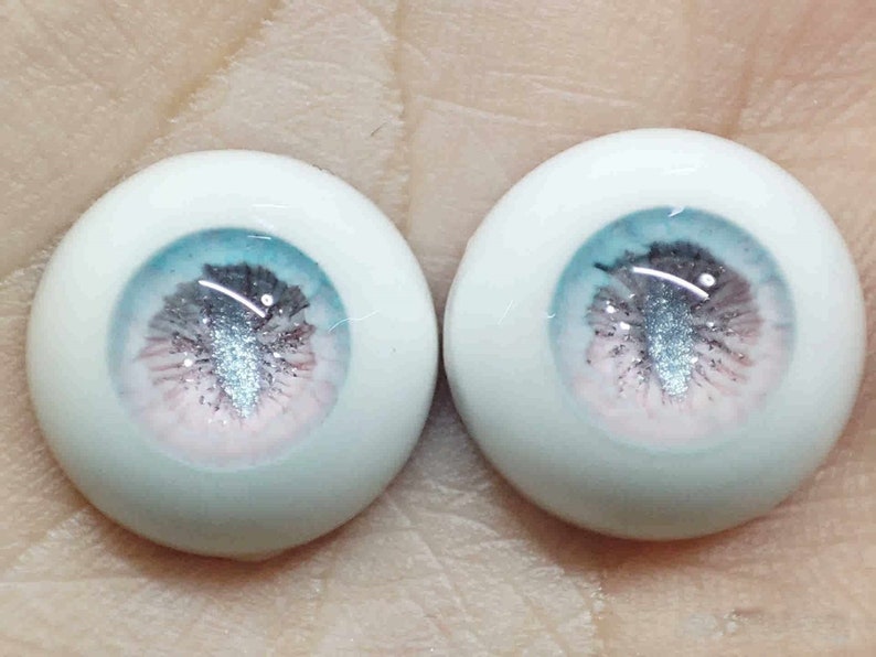 May include: Two white doll eyes with a blue, pink and silver glitter iris. The eyes are round and have a realistic appearance.