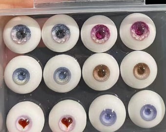 Puede incluir: Un conjunto de 12 ojos de muñeca en un contenedor de plástico transparente. Los ojos son de varios colores, incluyendo azul, marrón, rosa y morado. Algunos de los ojos tienen un diseño en forma de corazón.