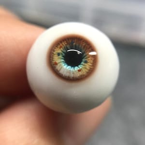以下が含まれることがあります： リアルな人形の目のクローズアップ。虹彩は茶色で青と黄色の斑点があり、瞳孔は黒で、強膜は白です。目は丸く、ガラスまたは同様の素材で作られているようです。