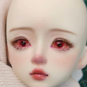 以下が含まれることがあります： 人形の顔のクローズアップ。肌は白く、頬はバラ色で、唇はふっくらとツヤがあります。人形は印象的な赤い目を持ち、まつげと眉毛が詳細に描かれています。全体的な美しさは繊細で芸術的です。