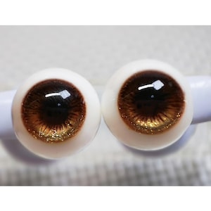 Könnte beinhalten: Ein Paar braune Glaspupillen mit goldenen Glitzerakzenten. Die Augen sind in einer weißen Plastikbasis befestigt.