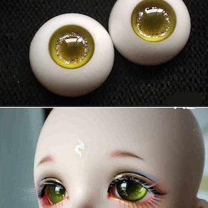 May include: A pair of green and gold doll eyes with a white iris and a glitter accent. The eyes are on a white background.