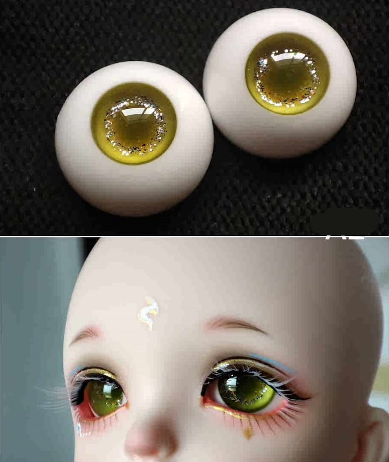 May include: A pair of green and gold doll eyes with a white iris and a glitter accent. The eyes are on a white background.