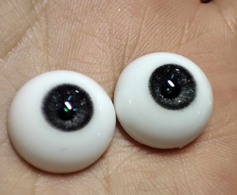 May include: Two realistic-looking glass doll eyes with a dark grey iris and a green fleck. The eyes are set against a light brown background.