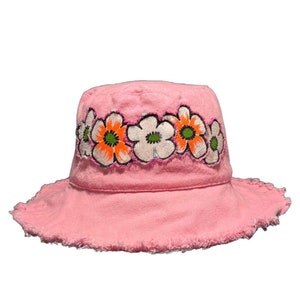 May include: A pink bucket hat with a frayed edge and a band of embroidered white flowers with orange and green centers.