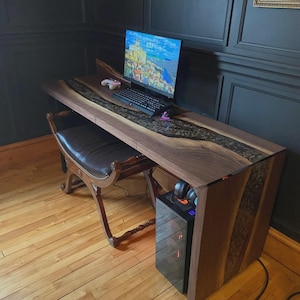 Op de afbeelding: Een op maat gemaakte houten bureau met een donkere walnoot afwerking en een heldere epoxy rivier. Het bureau heeft een computermonitor, toetsenbord en een gameconsole controller. Een computerkast en een leren stoel zijn ook aanwezig.