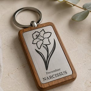 May include: A rectangular wooden keychain with a silver metal plate featuring a black line drawing of a daffodil. The text "December NARCISSUS" is below the flower. The keychain has a silver metal ring.