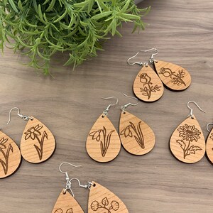 May include: Wooden teardrop earrings with engraved floral designs. The earrings feature various flower illustrations, including roses, daffodils, and chrysanthemums. Each earring has a silver-colored hook. The earrings are displayed on a wooden surface.