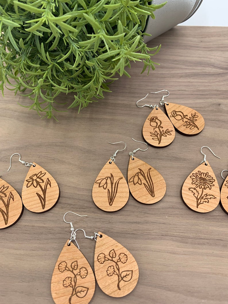 May include: Wooden teardrop earrings with engraved floral designs. The earrings feature various flower illustrations, including roses, daffodils, and chrysanthemums. Each earring has a silver-colored hook. The earrings are displayed on a wooden surface.