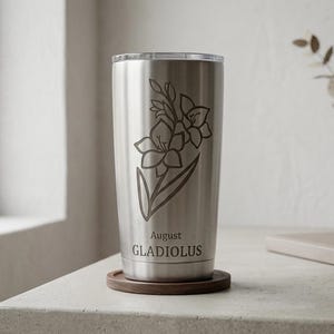 May include: A stainless steel tumbler with a floral design of a gladiolus flower. The text "August GLADIOLUS" is etched below the flower. The tumbler sits on a wooden coaster. A vase with a plant is in the background.
