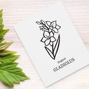 May include: A white card with a black line drawing of a gladiolus flower, labeled "August GLADIOLUS." The card is on a light wood surface, with green leaves in the background.