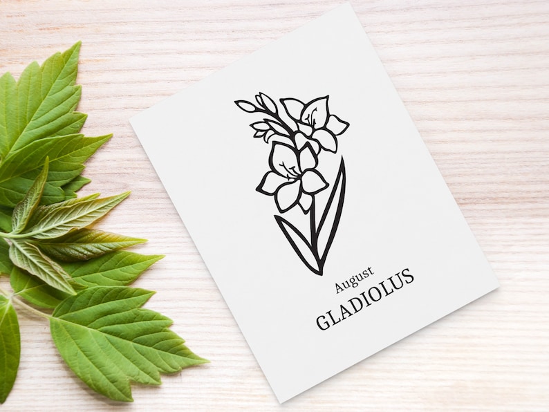 May include: A white card with a black line drawing of a gladiolus flower, labeled "August GLADIOLUS." The card is on a light wood surface, with green leaves in the background.