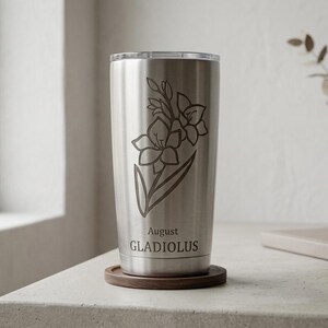 May include: A stainless steel tumbler with a floral design of a gladiolus flower. The text "August GLADIOLUS" is etched below the flower. The tumbler sits on a wooden coaster. A vase with a plant is in the background.