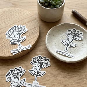 May include: A collection of honeysuckle stickers with a black and white floral design. One sticker is on a wooden coaster, another on a small ceramic dish, and others are scattered on a wooden surface. The stickers have the text "June HONEYSUCKLE".