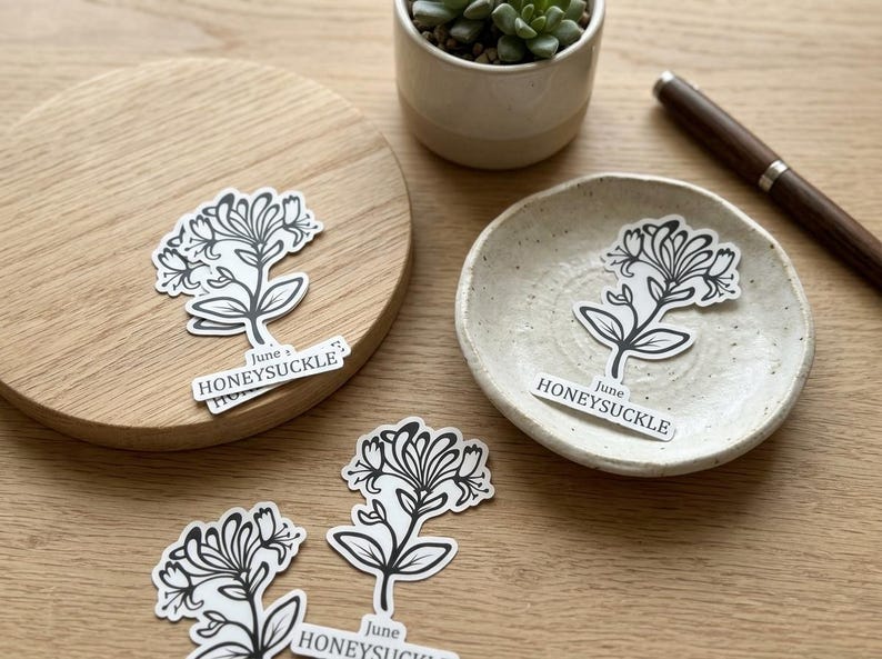 May include: A collection of honeysuckle stickers with a black and white floral design. One sticker is on a wooden coaster, another on a small ceramic dish, and others are scattered on a wooden surface. The stickers have the text "June HONEYSUCKLE".