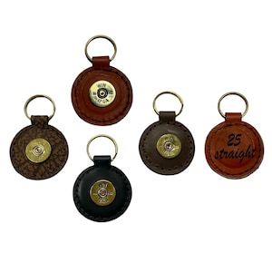 May include: Five leather keychains with brass key rings. The keychains are round with a brass bullet casing in the center. One keychain has the text "25 straight" engraved on it. The leather colors include brown, black, and tan.
