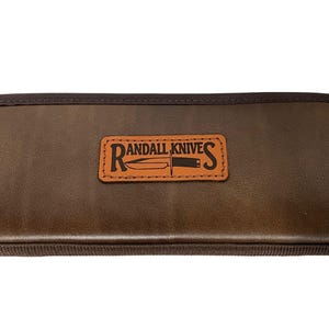 May include: A brown leather knife case with the words "Randall Knives" in a rectangular patch. The case is a dark brown color and has a textured appearance. The logo features a knife illustration.