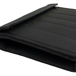 May include: Black, rectangular padded case with a flap. The case is made of a textured fabric and features several parallel, stitched lines across its surface. The edges are neatly finished with a contrasting trim.