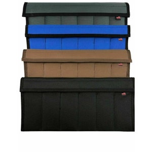 May include: A stack of rectangular, fabric pouches in various colors: dark green, blue, tan, and black. Each pouch has a flap closure and a small red and white tag. The pouches are designed with vertical stitching.