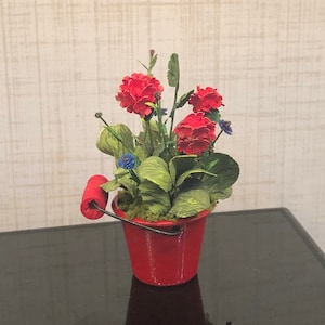 May include: A red metal bucket with a wooden handle, filled with a red and green floral arrangement. The flowers are red and blue.