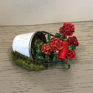 May include: A miniature white metal bucket with a red handle, filled with a red and orange flower arrangement. The bucket is sitting on a bed of green moss.