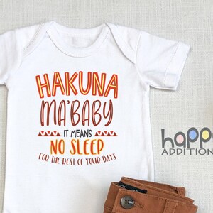 May include: A white baby onesie with the text "Hakuna Ma'baby It Means No Sleep For The Rest Of Your Days" in orange and red lettering. The onesie is laid out on a beige surface with brown trousers, brown shoes, a wooden rattle, and a cotton boll.