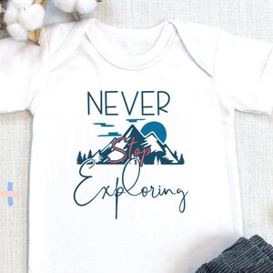 Puede incluir: Un body blanco para bebé con el texto "Never Stop Exploring" y un gráfico de una cordillera, un sol y árboles. El body se combina con pantalones grises a rayas.