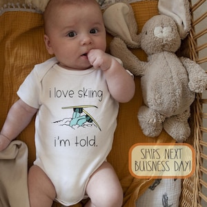Könnte beinhalten: Ein Baby in einem weißen Body mit dem Text "I love skiing, I'm told" und einer Skifahrer-Grafik. Das Baby liegt in einer Weidenkorb-Wiege mit einem Stoffhasen und einer Decke.
