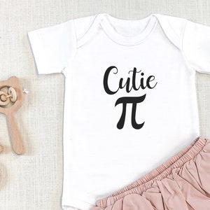 May include: White baby onesie with black text that reads "Cutie Pi". The onesie is paired with a pink ruffled diaper cover.