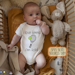 May include: A white baby onesie with a black and white illustration of a tennis racket and ball. The onesie says "I love tennis I'm told."