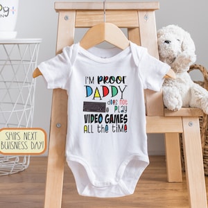 May include: White baby grow with a colourful graphic that reads "I'm Proof Daddy Does Not Play Video Games All The Time". The graphic includes a keyboard and mouse.