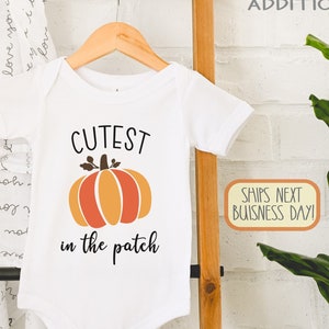 Könnte beinhalten: Weißer Baby-Strampler mit dem Text "CUTEST in the patch" und einem Cartoon-Kürbis in Orange und Gelb.