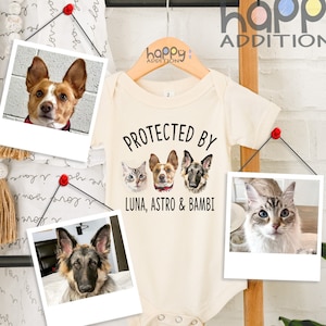 May include: A white baby onesie with the text "Protected by Luna, Astro & Bambi" and three pet portraits. The onesie is hanging on a wooden ladder with a white background.