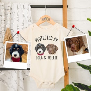 Puede incluir: Un body blanco para bebé con el texto "PROTECTED BY LOLA & MILLIE" impreso en la parte delantera. Dos fotos de perros se muestran encima del texto. El body está colgado en una escalera de madera.